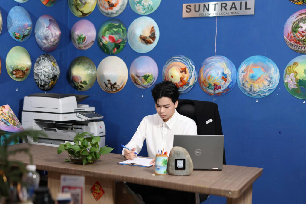 Vo Gia Kiet, Director of Suntrail Travel, at his desk in front of hand-painted conical hats at the Suntrail office